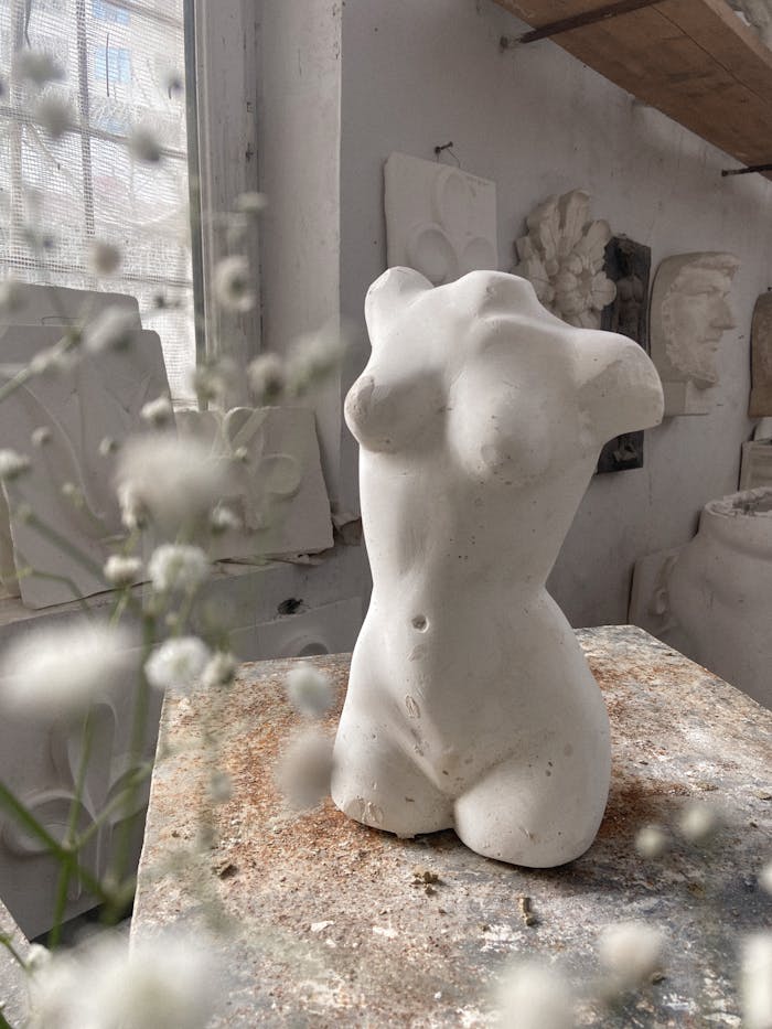 White clay sculpture of female torso placed on messy wooden desk in modern workshop with decorations and branch of plant